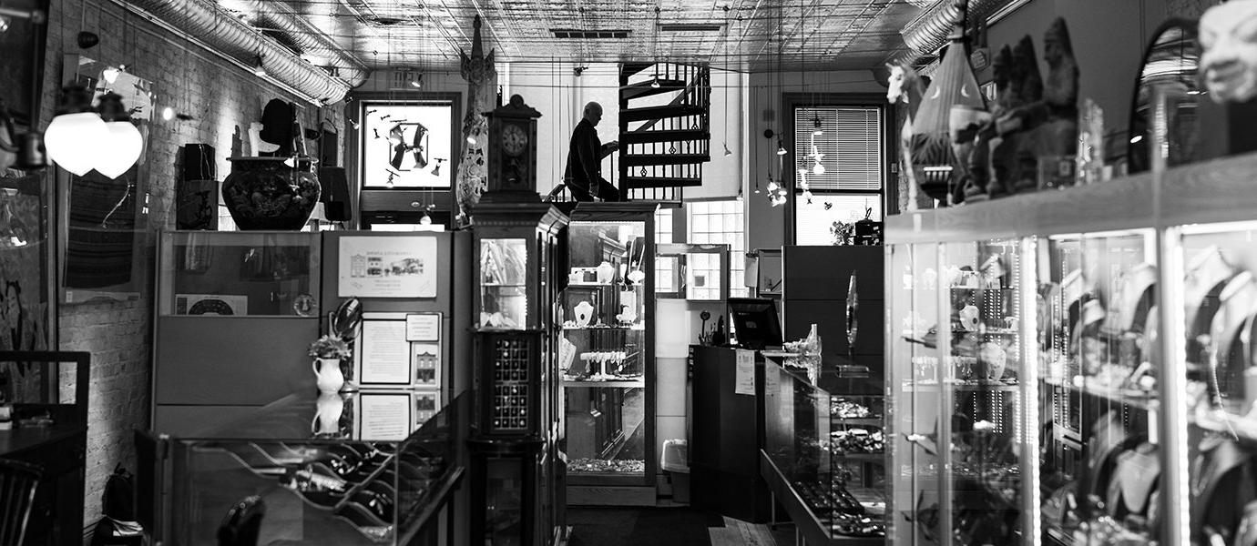 A black and white view down a jewelry store with a silhouette of a man walking up a winding staircase 